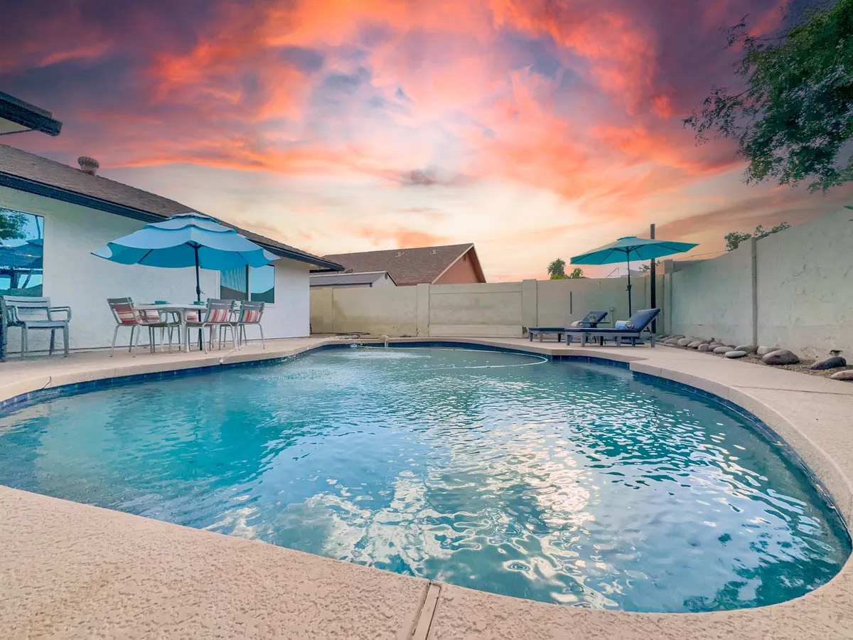 A tranquil backyard scene featuring a sparkling swimming pool, inviting patio furniture shaded by umbrellas, and an expansive sky above. The space is ideal for swimming, sunbathing, dining al fresco, or simply relaxing with family.