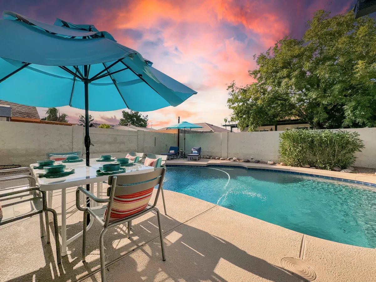 A vibrant backyard scene showcasing a sparkling swimming pool surrounded by lush greenery, a dining table set for alfresco meals, sun loungers beneath umbrellas, and the warm glow of sunset. The atmosphere is inviting and lively, perfect for both daytime fun and evening gatherings.
