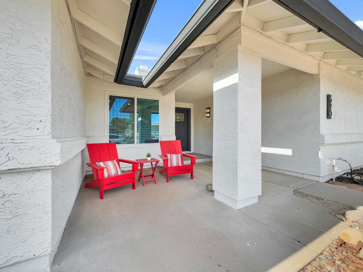 A cozy front porch scene featuring two red chairs and a small table, suggesting a relaxed atmosphere ideal for morning coffee, reading, or catching up with friends. The area is shaded and welcoming, offering a connection between the home and its neighborhood surroundings.