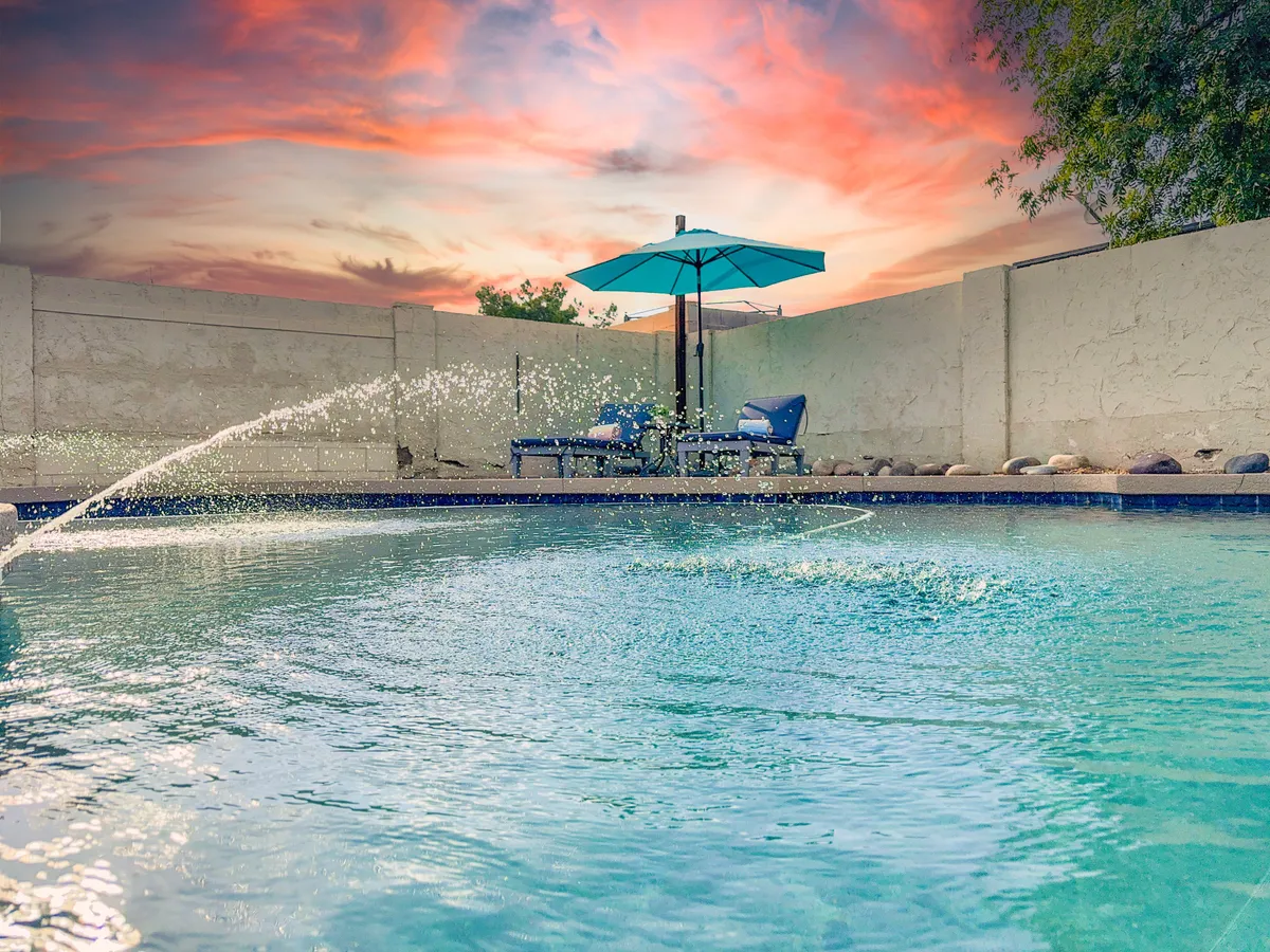 A tranquil poolside view with a swimming pool, umbrella-shaded lounge chairs, and a water feature. The serene mood suggests sunbathing, relaxing swims, and peaceful evening gatherings, with the sunset reflecting on the water.