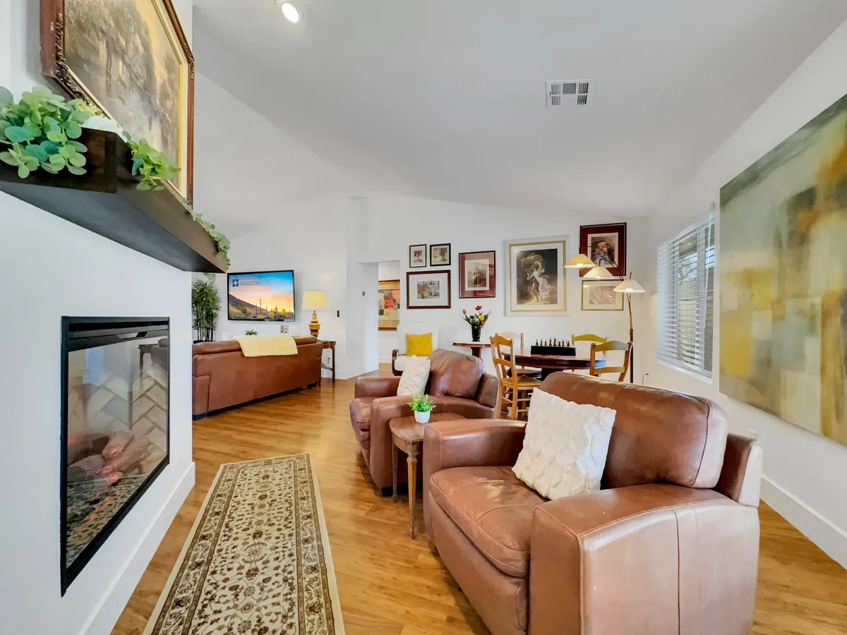 A spacious open-concept living and dining area, featuring a cozy fireplace as the focal point, stylish artwork on the walls, plush seating, and a connected dining space with a table ready for gathering. The room is awash in natural light, with smooth visual transitions between the lounging and dining areas.