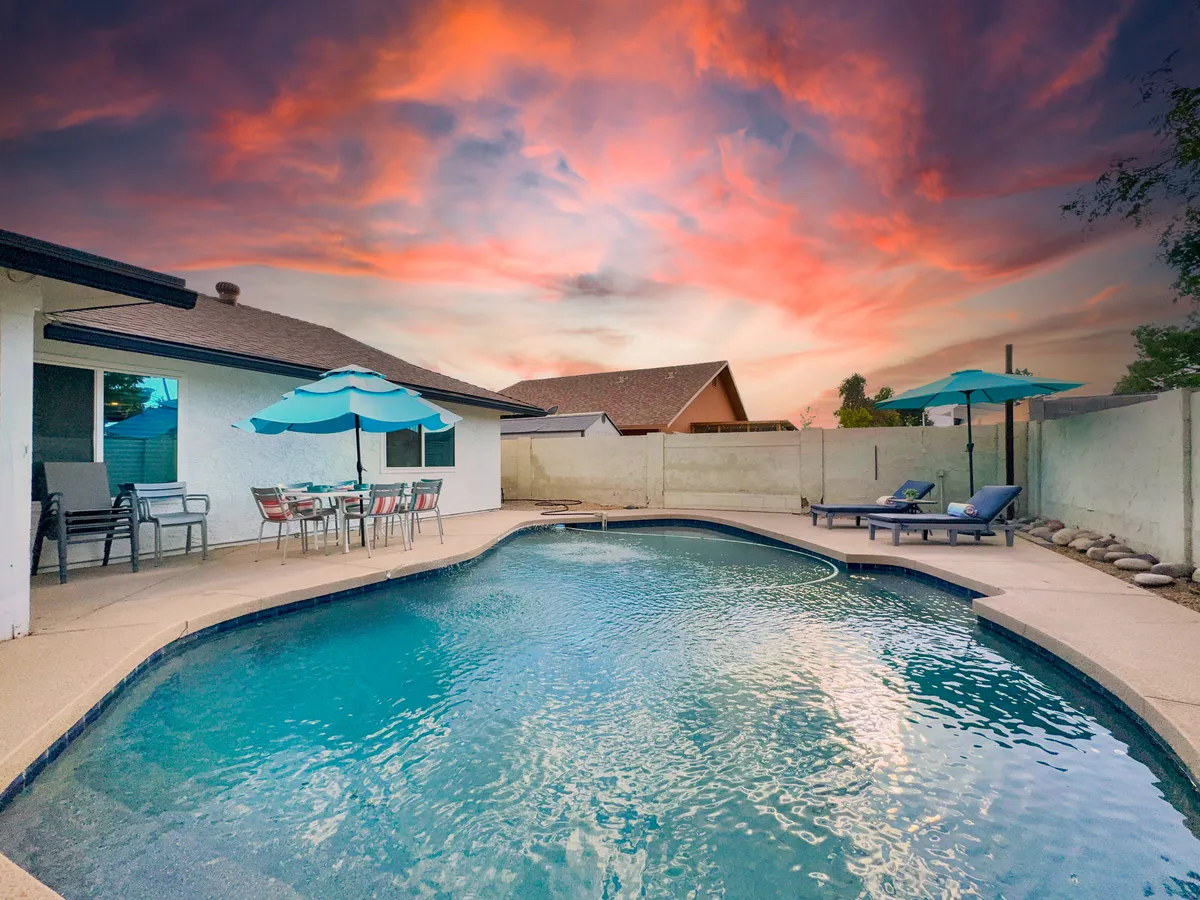 A sparkling swimming pool sits at the heart of an inviting backyard, surrounded by a cozy outdoor seating area with colorful umbrellas. The scene is set at sunset, with the sky glowing in soft hues, creating a cheerful and relaxing ambiance perfect for families to unwind or play together after a day of adventure.