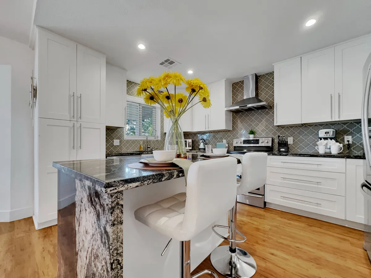 A bright, contemporary kitchen featuring gleaming granite countertops and sleek white cabinetry, accented with a vase of yellow flowers. Stainless steel appliances are prominently displayed, and bar stools along the central island invite casual dining or conversation.