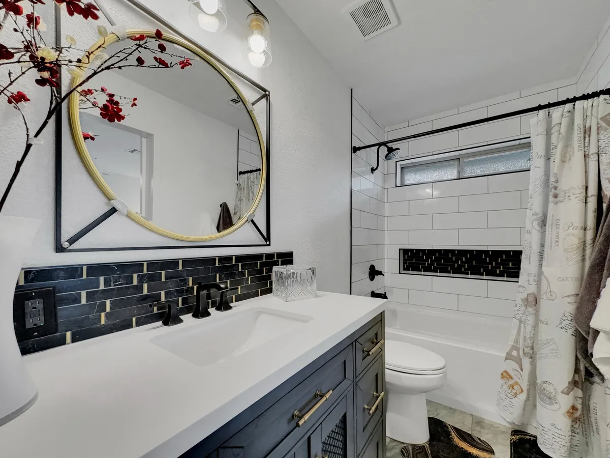 A modern bathroom with a large round mirror, stylish black and gold sink faucet, a decorative floral branch, a textured black backsplash, and a bathtub with a patterned shower curtain. The space is elegantly appointed and feels spa-like.