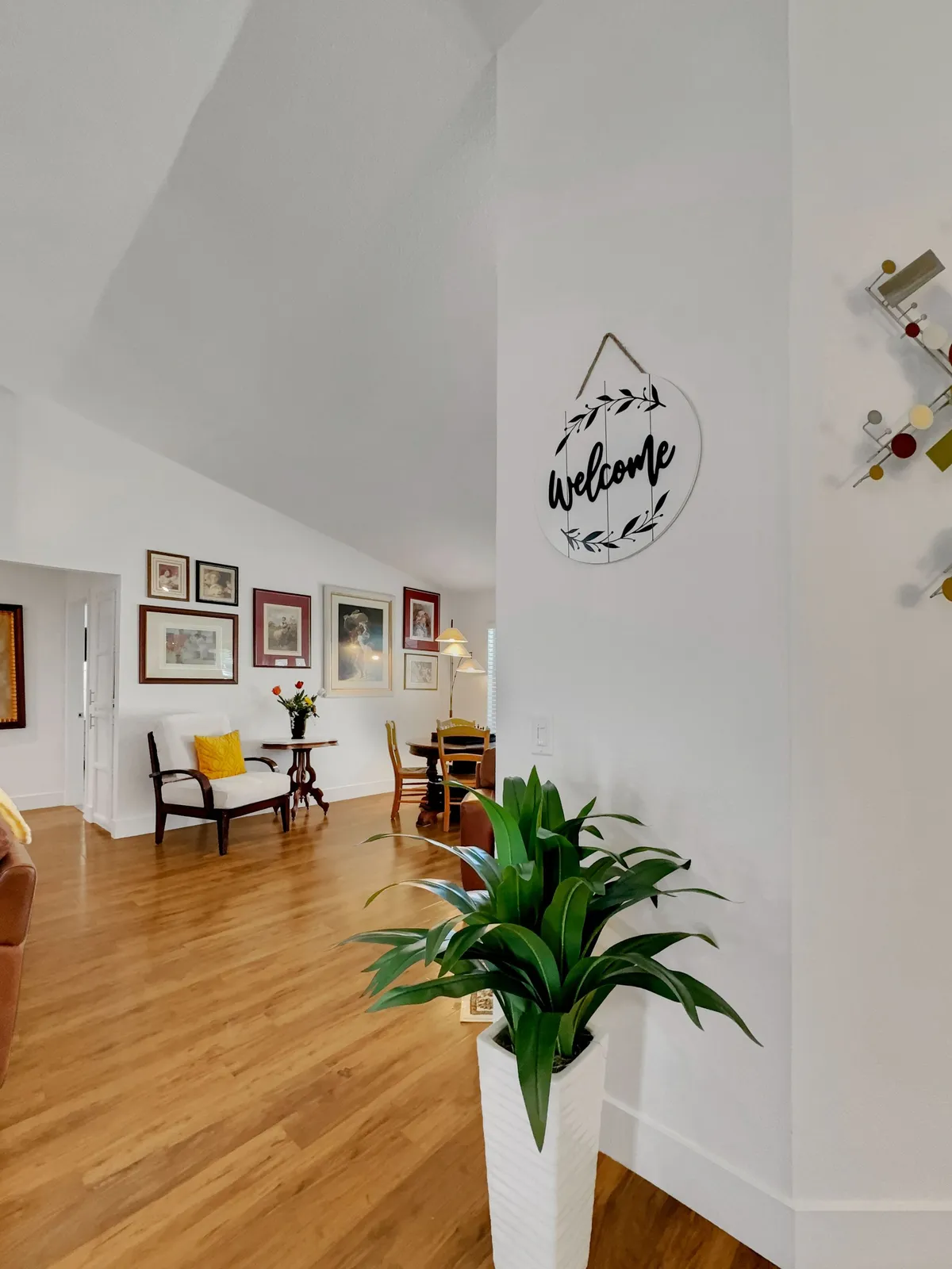 A bright, welcoming interior with a 'Welcome' sign, lush indoor plants, colorful artwork, and a comfortable seating area. The space is designed for gatherings, offering a cheerful and homely ambiance for creating shared memories.